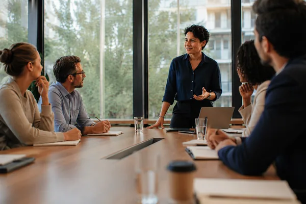 A woman standing at the head of a meeting table, speaking with confidence as colleagues listen