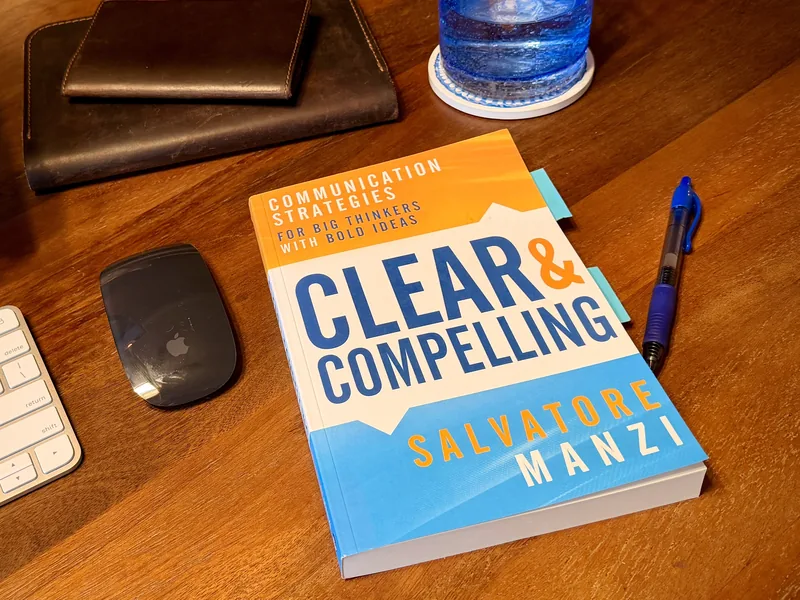 Clear and Compelling by Salvatore Manzi on a wooden desk beside a pen, a keyboard, and a mouse